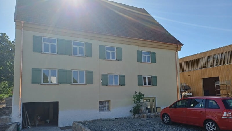 Ein zweistöckiges gelbes Haus mit grünen Fensterläden, einem Satteldach und einem roten Auto, das an einem sonnigen Tag davor geparkt ist.