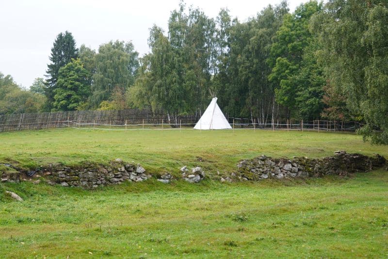 Ein weißes Tipi steht auf einer Wiese, umgeben von einem Holzzaun und Bäumen, mit einer niedrigen Steinmauer im Vordergrund - ein einladender Ort, der sich perfekt für Coaching-Sitzungen im Freien eignet.