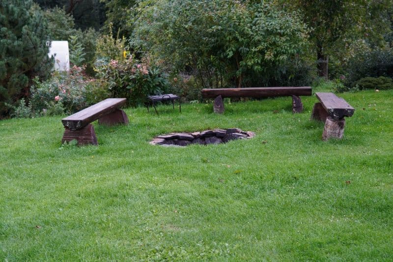 Drei Holzbänke umgeben eine einfache Feuerstelle aus Stein auf einem grasbewachsenen Rasen und schaffen einen einladenden Raum für Coaching-Sitzungen inmitten von Bäumen und Büschen im Hintergrund.
