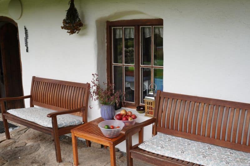 Zwei Holzbänke und ein kleiner Tisch mit Obstschalen bilden ein gemütliches Plätzchen für das Outdoor-Coaching, das vor einem weißen Stuckhaus mit Fenster und hängenden getrockneten Kräutern stattfindet.