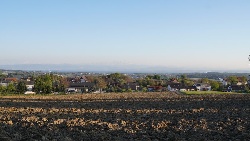 Ein gepflügtes Feld im Vordergrund mit Häusern und Bäumen im Hintergrund unter einem klaren blauen Himmel. Am Horizont sind in der Ferne die Berge zu erkennen.
