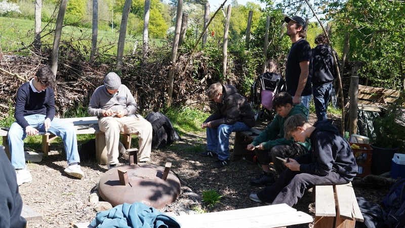 Eine Gruppe von Menschen sitzt auf Bänken und Holzstämmen um eine Feuerstelle im Freien, einige schauen auf Geräte herab, im Hintergrund sind Bäume und ein Holzzaun zu sehen.
