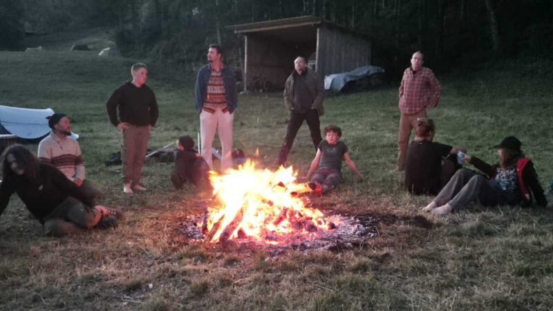 Eine Gruppe von Menschen sitzt und steht in der Abenddämmerung um ein großes Lagerfeuer auf einer grasbewachsenen Lichtung in der Nähe eines Holzschuppens.