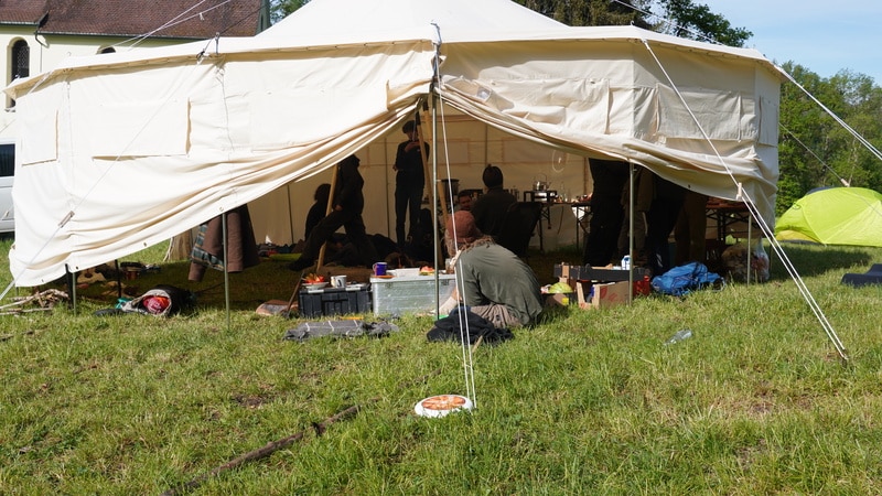 Mehrere Menschen sitzen und stehen innerhalb und außerhalb eines großen weißen Zeltes, das auf einer Wiese aufgebaut ist und in dem verschiedene Campingausrüstungen und Vorräte verstreut sind.