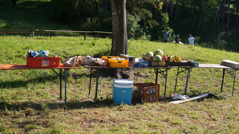 Eine Reihe von Klapptischen im Freien mit verschiedenen Lebensmitteln, Getränken und Vorräten; eine Kühlbox und Werkzeuge sind zu sehen, im Hintergrund Menschen und Bäume.