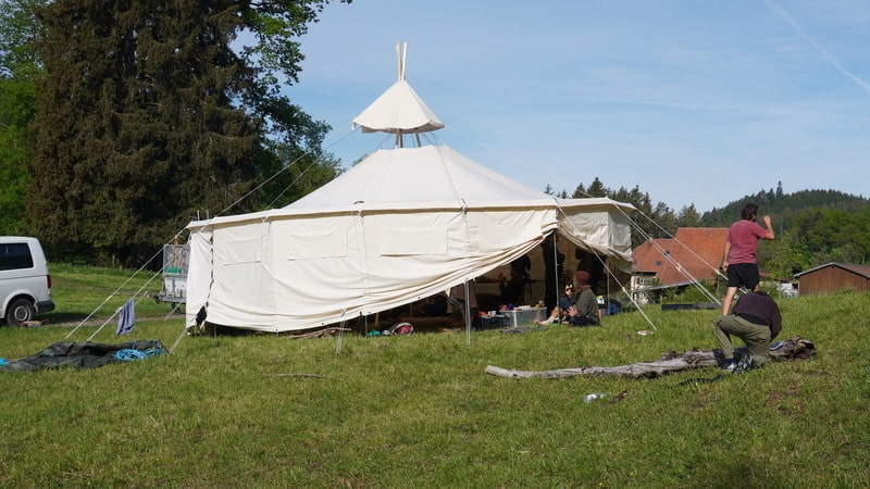 Eine Gruppe von Menschen hat ihr Lager um ein großes weißes Zelt auf einer Wiese aufgeschlagen, mit Campingausrüstung und einem weißen Lieferwagen in der Nähe.