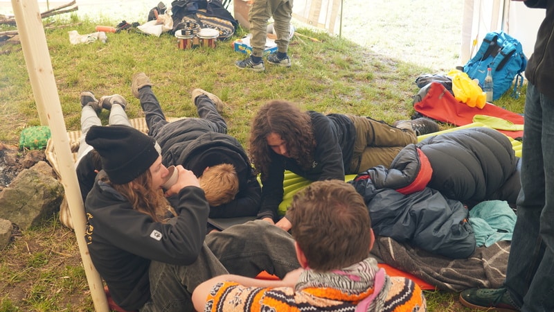 Mehrere Personen liegen auf Schlafmatten und Decken unter einem Vordach im Freien, umgeben von Rucksäcken und Campingausrüstung, mit Blick auf eine Grasfläche.